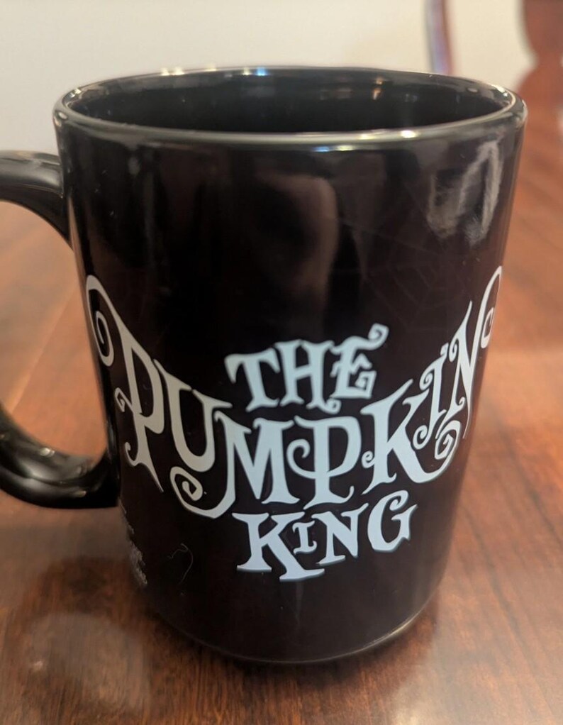 May include: A black ceramic mug featuring the text THE PUMPKIN KING printed in a stylised light blue font on the front. The mug has a smooth, glossy finish and a curved handle on the left side. A faint, subtle spiderweb pattern is visible in the background.