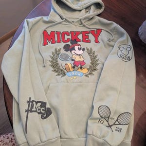 Puede incluir: Sudadera con capucha verde salvia con un gr&aacute;fico de Mickey Mouse jugando al tenis y la palabra "MICKEY" en rojo. La sudadera tiene un bolsillo de canguro y gr&aacute;ficos en las mangas, incluyendo raquetas de tenis cruzadas y el a&ntilde;o "1928".