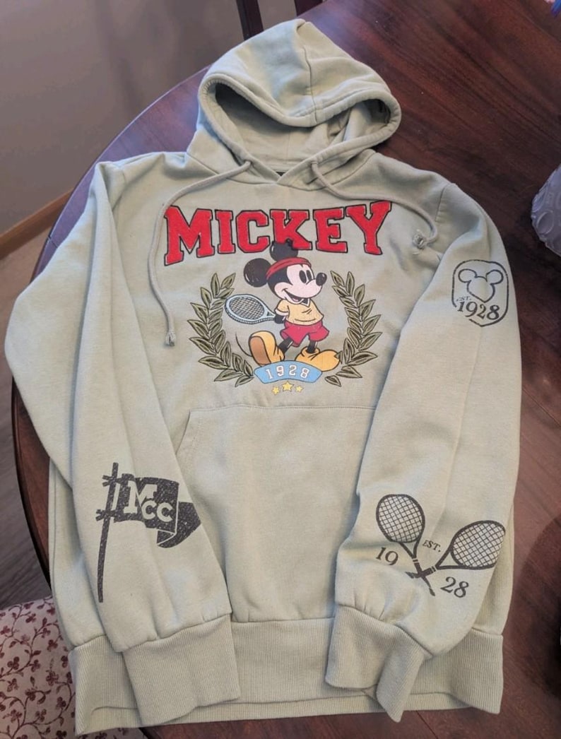 Puede incluir: Sudadera con capucha verde salvia con un gr&aacute;fico de Mickey Mouse jugando al tenis y la palabra "MICKEY" en rojo. La sudadera tiene un bolsillo de canguro y gr&aacute;ficos en las mangas, incluyendo raquetas de tenis cruzadas y el a&ntilde;o "1928".