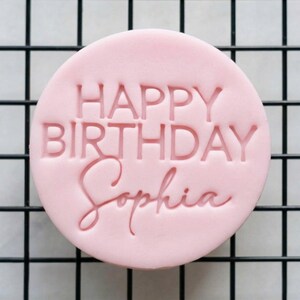Puede incluir: Un adorno para tarta redondo, rosa claro, con las palabras "HAPPY BIRTHDAY Sophia" en relieve. La fuente es sans-serif. El adorno está sobre un fondo de rejilla negra, creando un diseño simple y elegante.