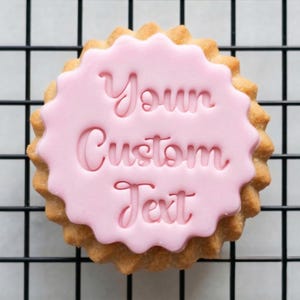 Może przedstawiać: Okrągłe ciastko z falistym brzegiem, pokryte różowym lukrem. Lukier zawiera napis "Your Custom Text" w kursywie. Ciastko leży na czarnym drucianym ruszcie.
