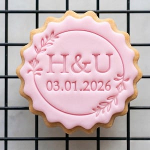 May include: A round biscuit with a pink icing design. The icing features the letters "H & U" and the date "03.01.2026" within a floral wreath. The biscuit has a scalloped edge and is set on a black wire rack.