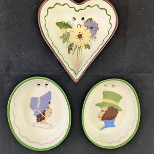 May include: A set of three ceramic dishes with two small holes at the top for hanging. One heart-shaped dish features a yellow and purple floral bouquet. Two oval dishes feature profile illustrations of figures wearing hats in blue and green.