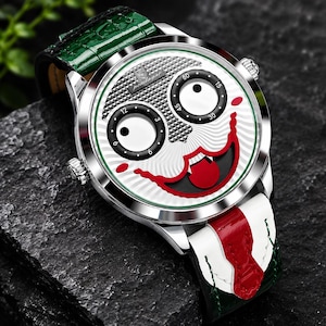 Puede incluir: Un reloj de pulsera plateado con una esfera blanca que presenta un dise&ntilde;o de rostro estilizado, dos subesferas circulares negras, un gr&aacute;fico de boca roja y el texto "Pondagar". El reloj tiene una correa de cuero verde y blanca con detalles rojos.