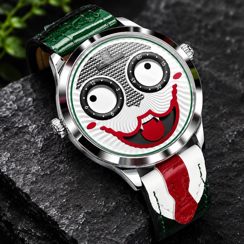Puede incluir: Un reloj de pulsera plateado con una esfera blanca que presenta un dise&ntilde;o de rostro estilizado, dos subesferas circulares negras, un gr&aacute;fico de boca roja y el texto "Pondagar". El reloj tiene una correa de cuero verde y blanca con detalles rojos.
