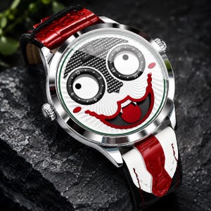 Puede incluir: Un reloj de pulsera con caja plateada y esfera blanca que presenta un dise&ntilde;o de rostro estilizado con dos subesferas circulares para los ojos, una boca roja y una lengua. El reloj tiene una correa de cuero roja y blanca con acabado texturizado.