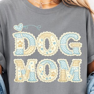 Puede incluir: Camiseta gris con las palabras "DOG MOM" en un diseño de patchwork. Las letras son azules y beige con detalles florales y de huellas de patas, y un borde festoneado. Un corazón azul y un detalle en espiral están encima de la palabra "DOG".