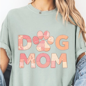 Puede incluir: Camiseta verde claro con las palabras "DOG MOM" en un diseño de patchwork. La "O" de DOG se reemplaza con una huella de pata. Las letras y la huella son en tonos rosas y naranjas.