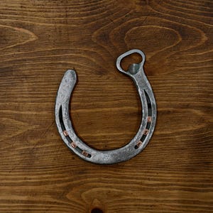 May include: A silver horseshoe bottle opener with copper-colored accents. The horseshoe is set against a rustic, wooden background. The bottle opener is a unique and functional item.