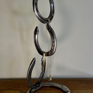 May include: A metal horseshoe jewelry stand. The stand is made of three connected horseshoes, with a base horseshoe. The stand is holding several necklaces and a bracelet. The stand is on a wooden surface.