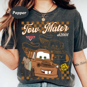 May include: Dark gray t-shirt featuring a cartoon image of Tow Mater from the Cars movie. The design includes the text "Tow Mater st2008" above the character, with a Route 66 sign and a checkered background.