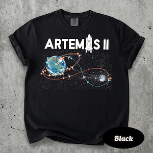 May include: Black t-shirt with the text "ARTEMIS II" and a graphic of Earth, a rocket, and a flight path. The design includes a depiction of the moon and stars. The shirt is made of a soft material.