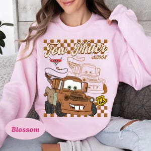 May include: A light pink sweatshirt featuring a graphic of Tow Mater from the Cars movie. The design includes the text "Tow Mater st.2008" and a Route 66 sign. The word "Blossom" is printed at the bottom.