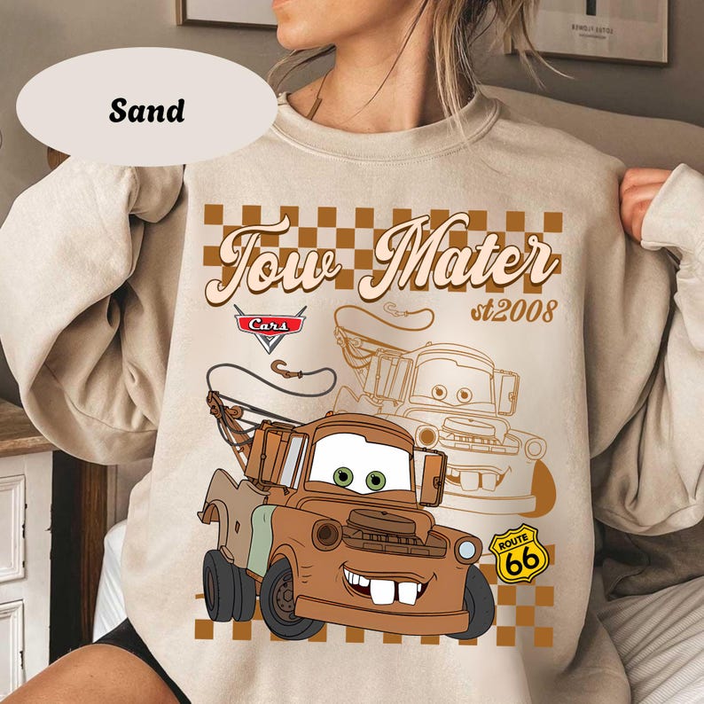 May include: Sand-colored sweatshirt featuring a graphic of Tow Mater from the Cars movie. The design includes the text "Tow Mater st 2008" and a Route 66 sign. The sweatshirt has a crew neck and long sleeves.