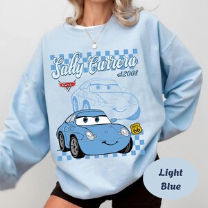 May include: Light blue sweatshirt featuring a cartoon image of Sally Carrera from the movie Cars. The design includes the text "Sally Carrera st.2008" and a Route 66 emblem. The sweatshirt is light blue.