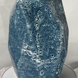 May include: A textured, blue ceramic vase with a unique, irregular shape. The vase has a speckled pattern with white accents, giving it a handcrafted appearance. The vase is sitting on a white plate.