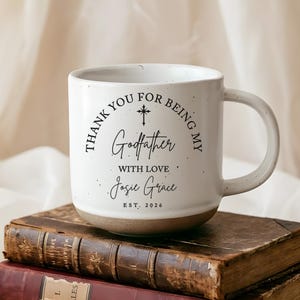 May include: A white ceramic mug with a speckled finish and a handle, featuring black printed text that reads Thank You For Being My Godfather With Love Josie Grace Est. 2026, accompanied by a small cross icon centered above the word Godfather.