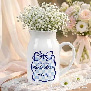 May include: A white ceramic pitcher filled with white baby's breath flowers. The front features a blue and white checkered square graphic with a bow on top, the text "It's giving Godmother" in blue script, a small heart, and the name "Emily".