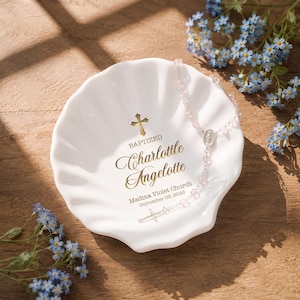 May include: A white shell-shaped ceramic dish featuring gold text that reads BAPTIZED Charlotte Angelotte Madina Violet Church September 09, 2026, with a gold cross icon above the name and a delicate rosary with light pink beads resting inside.