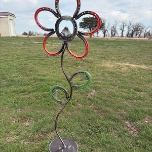 May include: A metal flower sculpture crafted from horseshoes. The flower's petals are formed from red and orange horseshoes, with a central ring of horseshoes. The stem and leaves are made of metal, with green horseshoes for the leaves. The sculpture is mounted on a round metal base.