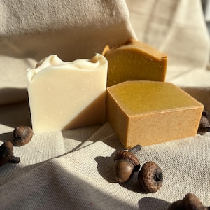 May include: Three bars of handmade soap in varying shades of beige and brown, with a few acorns scattered around. The soap bars have different shapes, with one having a wavy top. The soaps are on a beige linen cloth.