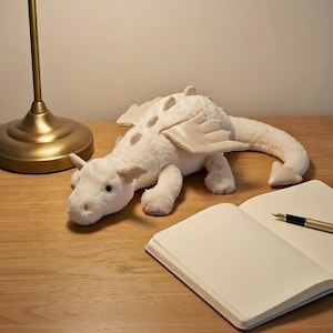 Puede incluir: Un peluche de dragón blanco con detalles beige, como alas y espinas dorsales. El dragón está sobre una superficie de madera, junto a un cuaderno abierto y un bolígrafo. El juguete mide aproximadamente 30 cm de largo.