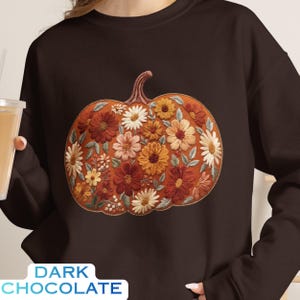 May include: A dark chocolate brown crewneck sweatshirt featuring a large, central graphic of a pumpkin filled with embroidered-style floral patterns in shades of orange, rust, yellow, and white, accented with small green leaves and stems.