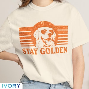 May include: An ivory short-sleeve t-shirt featuring a distressed orange graphic of a dog wearing sunglasses inside a circular frame with horizontal lines. The text "STAY GOLDEN" is printed in orange capital letters below the dog illustration.