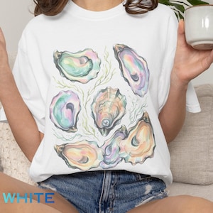 May include: White t-shirt featuring a watercolor illustration of several open oyster shells in pastel colors. The shells have iridescent hues of pink, green, and yellow, with a few pearls. The word "WHITE" is printed at the bottom.