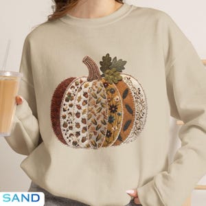 May include: A sand-colored crewneck sweatshirt featuring a large, central pumpkin graphic. The pumpkin is composed of vertical panels with diverse patterns, including acorns, mushrooms, corn, and floral motifs in shades of brown, gold, and green.