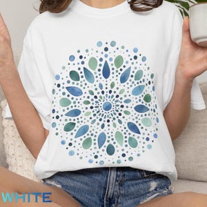 May include: White t-shirt featuring a watercolor mandala design in shades of blue and green. The mandala is composed of dots and teardrop shapes arranged in a circular pattern. The word "WHITE" is printed at the bottom of the shirt.