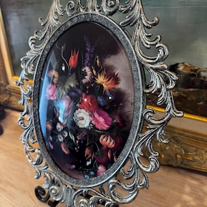 May include: Ornate silver-colored oval picture frame with a floral still life painting. The frame has a detailed, scrolling design and is supported by a black metal easel. The painting features a dark background with colorful flowers.