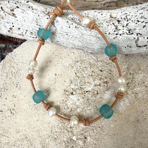 Peut inclure: Un bracelet composé d'un cordon en cuir beige enfilé avec une alternance de perles blanches et de perles en verre marin bleu clair translucide. Les perles sont maintenues par des nœuds sur le cordon, qui forme une fermeture à boucle et nœud.