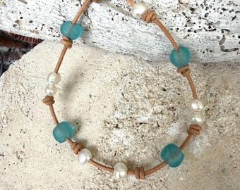 May include: A bracelet featuring a tan leather cord strung with alternating white pearls and translucent light blue sea glass beads. The beads are secured in place by small knots tied in the leather cord, which also forms a loop and knot closure.