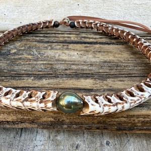 Może przedstawiać: Ręcznie robiony naszyjnik o wzorze przypominającym kość z brązowymi akcentami. Naszyjnik ma centralną, opalizującą, ciemnozieloną perłę. Naszyjnik jest prezentowany na zwietrzałej drewnianej powierzchni.
