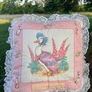 May include: A square decorative cushion featuring a central illustration of Jemima Puddle-Duck wearing a blue bonnet and shawl, surrounded by pink flowers. The cushion has a pink border, white lace trim, and the text JEMIMA PUDDLE-DUCK printed at the bottom.