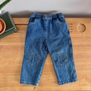 May include: Blue denim pants for toddlers, featuring an elastic waistband and heart-shaped patches on the knees. The pants are made of a medium-wash denim and have two front pockets.