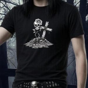 May include: Black t-shirt featuring a detailed graphic of a skull with a rose, a cross, and a bed of stones. The design is in grayscale, creating a gothic or alternative aesthetic. The shirt has short sleeves and a crew neck.