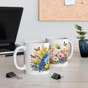 Op de afbeelding: Twee witte keramische mokken met bloemmotieven en de tekst "Be Kind". Eén mok heeft een boeket van gele, blauwe en paarse bloemen met oranje vlinders. De andere mok heeft een bij en kleurrijke bloemen.