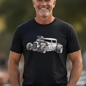May include: A black short-sleeve t-shirt featuring a detailed white graphic illustration of a classic hot rod car with a large exposed engine and flame decals on the side, paired with a dark baseball cap and blue denim jeans.