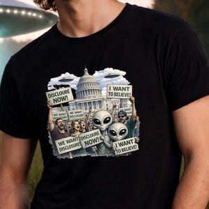 May include: Black t-shirt featuring a graphic of aliens protesting in front of a government building. The aliens hold signs that read "I WANT TO BELIEVE!" and "DISCLOSURE NOW!" with the text "WE WANT DISCLOSURE" on other signs.