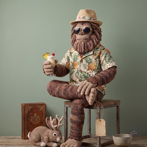 Peut inclure: Une créature brune crochetée avec une crinière hirsute, portant un fedora en paille avec un ruban floral, des lunettes d'aviateur et une chemise à manches courtes boutonnée avec un imprimé ananas. Elle tient une boisson crochetée avec ananas.