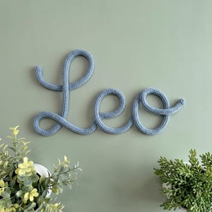 May include: A light blue knitted cord shaped into the cursive name Leo. The letters are formed from a single continuous piece of yarn with a visible textured stitch pattern, creating a soft and flexible appearance for this wall-mounted sign.