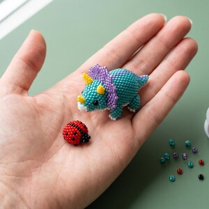 May include: A small triceratops figurine and a ladybug figurine crafted from tiny colorful seed beads. The triceratops is teal with a purple frill and yellow horns. The ladybug is red with black spots. A spool of fine seed beads is nearby.