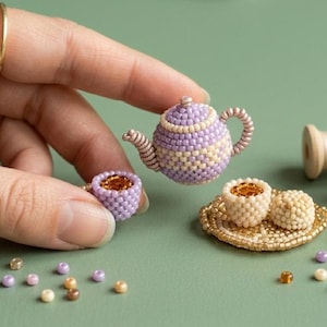 May include: A miniature teapot and two cups crafted from tiny lavender, cream, and gold beads. The teapot features a geometric pattern, while the cups are filled with amber-coloured beads. A gold beaded saucer and loose beads are scattered nearby.
