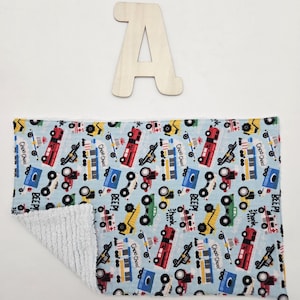 May include: A light blue baby blanket with a pattern of red, yellow, green, and blue vehicles. The blanket has the words "Choo Choo!" and "BEEP!" printed on it. A wooden letter "A" is above the blanket.