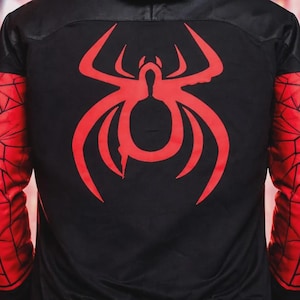 May include: Black bomber jacket with a red spider emblem on the back and red spiderweb-patterned sleeves. The jacket has a black hood and red trim at the waist and cuffs. The background is a blurred city scene.