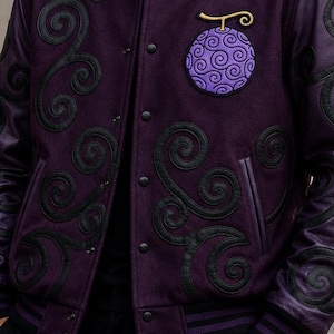 Puede incluir: Chaqueta universitaria morada con detalles negros y un parche circular con un diseño de fruta morada. La chaqueta tiene patrones de remolinos negros y un cuello negro. La chaqueta se usa con pantalones negros.