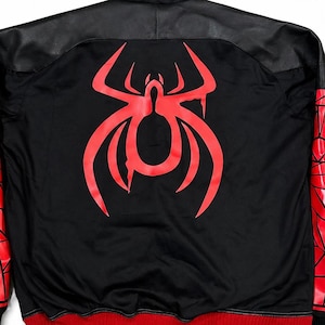 May include: Black hooded jacket with a red spider emblem on the back. The jacket features red and black geometric designs on the sleeves, a black hood, and a red trim at the bottom. The jacket has a black leather-like panel across the shoulders.