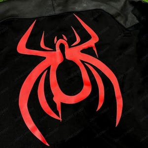 May include: Black jacket with a red spider emblem on the back. The jacket has a hood and a leather-like panel across the shoulders. The spider design is a bold, stylized graphic.